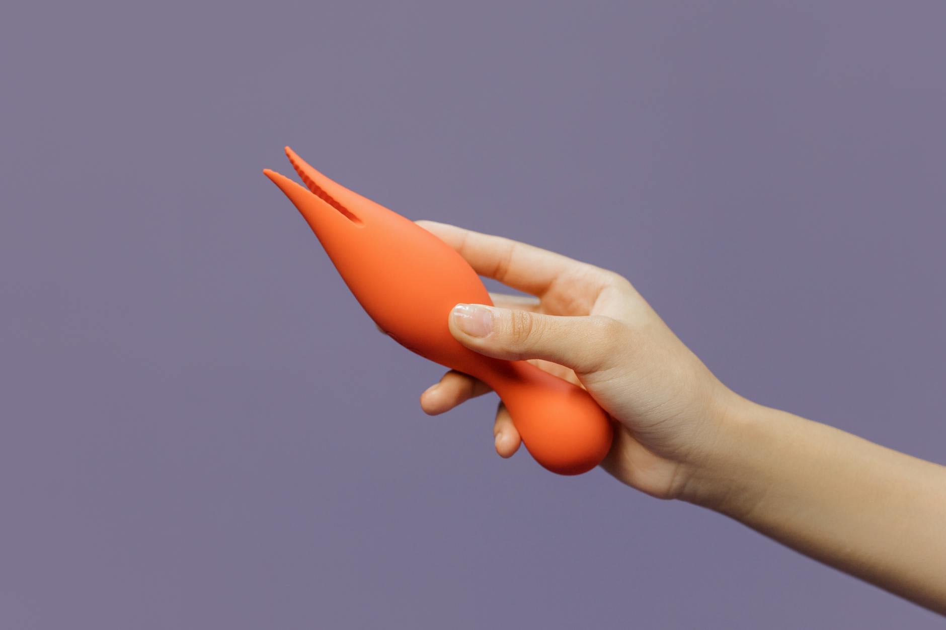 Eine Hand hält einen orangefarbenen Vibrator vor minimalistische violette Kulisse, modern und sinnlich.